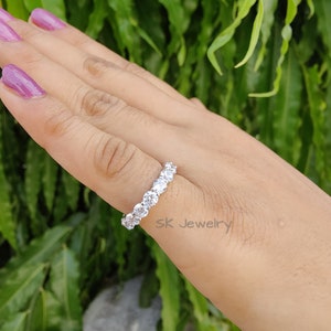 May include: A silver ring with a row of clear round stones. The ring is on a hand with pink nail polish. The background is green foliage.