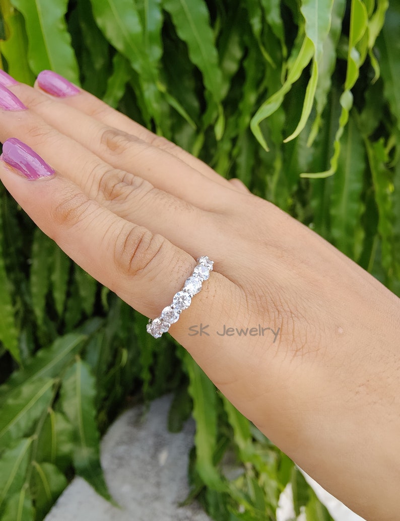 May include: A silver ring with a row of clear round stones. The ring is on a hand with pink nail polish. The background is green foliage.