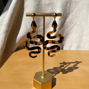 May include: A pair of gold tone snake-shaped earrings with a tortoiseshell pattern. The earrings are displayed on a gold stand.