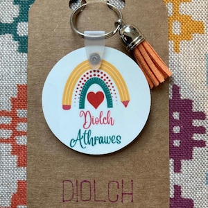 May include: A round keychain featuring a rainbow and heart design, with the words "Diolch Athrawes". It has a silver ring and an orange tassel. The keychain is attached to a brown tag with the text "Diolch Crefftiau Ty Celyn Crafts".