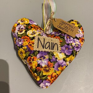 Può includere: Ornamento in tessuto a forma di cuore con un motivo floreale giallo, viola e arancione. La parola "Nain" è cucita su una toppa di iuta. Una piccola etichetta recita "Crefftau Ty Celyn Crafts."