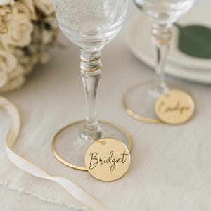 May include: Two clear wine glasses with gold rims and stems. A gold circular tag with the name "Bridget" in black script hangs from the base of one glass. A second glass has a tag with the name "Caitlyn". A white ribbon is draped across the table.