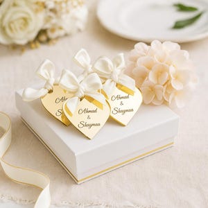 Può includere: Una scatola regalo bianca con finiture dorate, sormontata da tre etichette dorate a forma di cuore legate con fiocchi di raso bianco. Ogni etichetta riporta i nomi "Ahmad & Shaymaa". Composizioni floreali e nastri completano la presentazione.