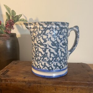 May include: A vintage stoneware pitcher with a blue and white speckled pattern. The pitcher has a spout and a handle. A blue stripe accents the base. The pitcher is sitting on a wooden box.