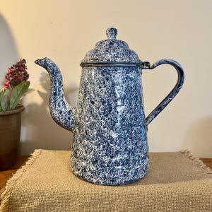 May include: Vintage blue and white speckled enamelware coffee pot. The pot has a tall, slender shape with a curved spout and a handle. The lid is also speckled and has a small knob. The pot is sitting on a tan burlap cloth.