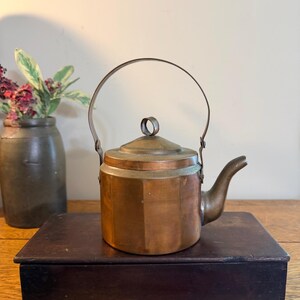 May include: A copper teapot with a hexagonal body, a curved spout, and a rounded lid featuring a small circular handle. A thin, arched metal handle is attached to the sides of the teapot, which has a warm, metallic finish and visible seams.