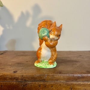 May include: A ceramic squirrel figurine with a reddish-brown coat and a white belly, holding a green sphere. The squirrel is perched on a patch of green grass. The figurine is a decorative item.