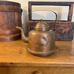 May include: A vintage copper teapot with a spout, handle, and lid. The teapot sits on a small, round base. The copper has a warm, aged patina. The background includes a wooden barrel and a rustic wooden box.
