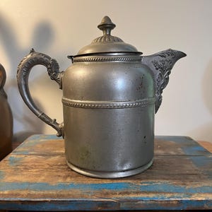 May include: Antique pewter teapot with ornate handle and spout details. The teapot has a domed lid with a finial and is set on a wooden surface with blue and brown paint. The teapot is a vintage item.