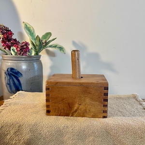 May include: A wooden butter mold with a rectangular box shape and a cylindrical handle. The box is made of light-colored wood with visible joinery. The butter mold sits on a tan burlap cloth.