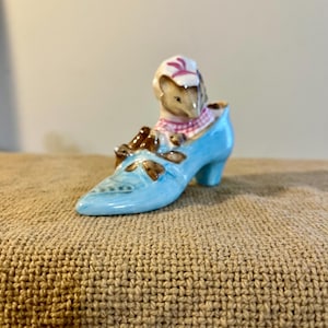May include: A ceramic figurine of a mouse wearing a pink and white checkered scarf and hat, sitting in a light blue shoe. The shoe has gold accents and a small heel. The figurine is on a textured brown surface.