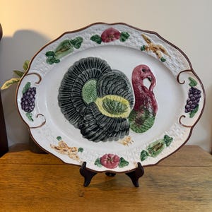 May include: An oval ceramic serving platter featuring a raised, three-dimensional turkey in the center. The rim is decorated with a white basket-weave texture and painted relief motifs of grapes, carrots, and tomatoes in green, purple, and red.