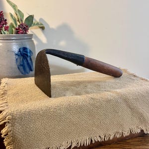 May include: A vintage garden hoe with a dark metal blade and a wooden handle. The handle is attached to the blade with a black metal collar. The hoe is resting on a tan burlap cloth with a fringed edge. A stoneware pot with a blue floral design is in the background.