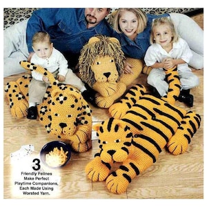 May include: Three crocheted animal floor cushions: a yellow and black striped tiger, a yellow and brown spotted leopard, and a yellow lion with a brown mane. The cushions are on a wood floor. The text "3 Friendly Felines Make Perfect Playtime Companions, Each Made Using Worsted Yarn." is at the bottom of the image.
