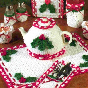 Puede incluir: Funda de tetera de ganchillo blanco con detalles de acebo rojo y verde. La funda está sobre un mantel individual de ganchillo blanco con detalles de acebo rojo y verde. También hay fundas de ganchillo para los saleros y pimenteros, un portavelas y un portacubiertos.