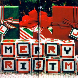 May include: A set of 10 white fabric blocks with red and black trim spelling out "Merry Christmas". The blocks are arranged in a row and are sitting in front of three wrapped gifts with red and green ribbon.