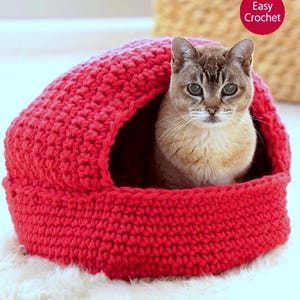 May include: A red, crocheted cat bed with a round opening. A gray and tan cat sits inside the bed, looking directly at the camera. The words "Easy Crochet" are in a red circle in the upper right corner.