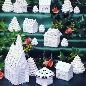 Könnte beinhalten: Eine Anordnung weiß gehäkelter Miniaturhäuser und Weihnachtsbäume auf einer grünen Oberfläche. Die Häuser haben unterschiedliche Designs, einige mit Satteldächern und andere mit Flachdächern. Rosa Rosen und grünes Laub schmücken die Szene.