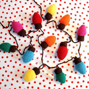 May include: A string of crocheted light bulbs in various colors, including red, orange, yellow, green, blue, and pink, hanging on a white background with red polka dots.
