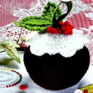 May include: A crocheted Christmas pudding ornament with a white icing top, red berries, and green leaves.