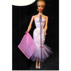 May include: A vintage Barbie doll wearing a white lace dress with a tulle skirt and a pink scarf. She is holding a microphone and standing on a clear stand.