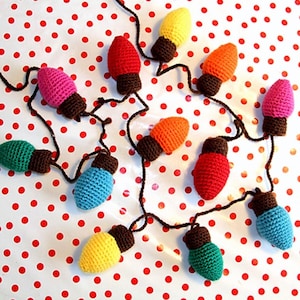 Puede incluir: Una guirnalda de luces navideñas tejidas a ganchillo en varios colores. Las luces son rojas, amarillas, naranjas, rosas, verdes y azules, con bases marrones. Están conectadas por un cable negro y dispuestas sobre una superficie blanca con lunares rojos.