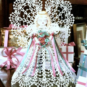 May include: White crocheted angel ornament with a lace dress, wings, and a halo. The angel is holding a bouquet of pink and green flowers and is surrounded by wrapped gifts.