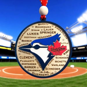 May include: A wooden Toronto Blue Jays ornament featuring a blue jay logo, player names, and "World Series 2025" text. The round ornament has a red maple leaf and a red and white ribbon. The background shows a baseball field.