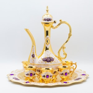 May include: A gold-plated tea set with a decorative white and purple floral design. The set includes a teapot, six cups, and a tray.