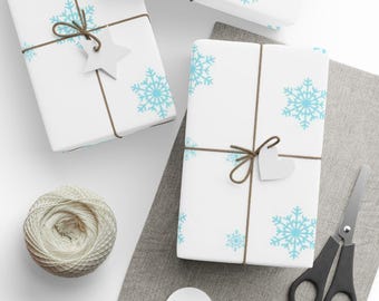 May include: White gift boxes wrapped in paper with light blue snowflake patterns, tied with brown twine and adorned with star and heart-shaped tags. A ball of twine, scissors, and blank tags are also present, suggesting a holiday or winter theme.