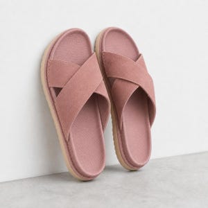 Op de afbeelding: Een paar oudroze slippers met brede, gekruiste banden over de bovenkant. Het voetbed heeft een gestructureerd oppervlak en de zool is lichtbruin van kleur. De slippers staan rechtop tegen een verticaal oppervlak aan.