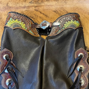 May include: A pair of brown leather chaps with sunflower embroidery and fringe detail. The chaps have a silver buckle and are decorated with silver conchos.