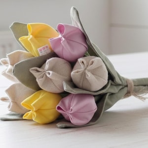 Op de afbeelding: Een boeket van handgemaakte stoffen tulpen in de kleuren geel, zachtroze en beige. De bloemen hebben gestikte bloemblaadjes en groene stoffen bladeren, die allemaal zijn samengebonden met een eenvoudig touwtje aan de basis van de stelen.