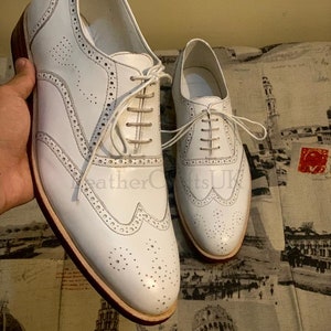 Puede incluir: Un par de zapatos de vestir de cuero blanco con dise&ntilde;o de punta de ala y cierre de cordones. Los zapatos tienen una suela de cuero marr&oacute;n y una parte superior de cuero blanco.