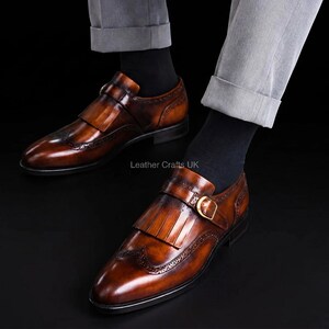 May include: A pair of burnished tan leather monk strap shoes featuring a kiltie fringe detail, a gold-tone buckle, and decorative brogue perforations along the toe cap. The shoes are worn with black socks and light gray trousers. Text reads Leather Crafts UK.