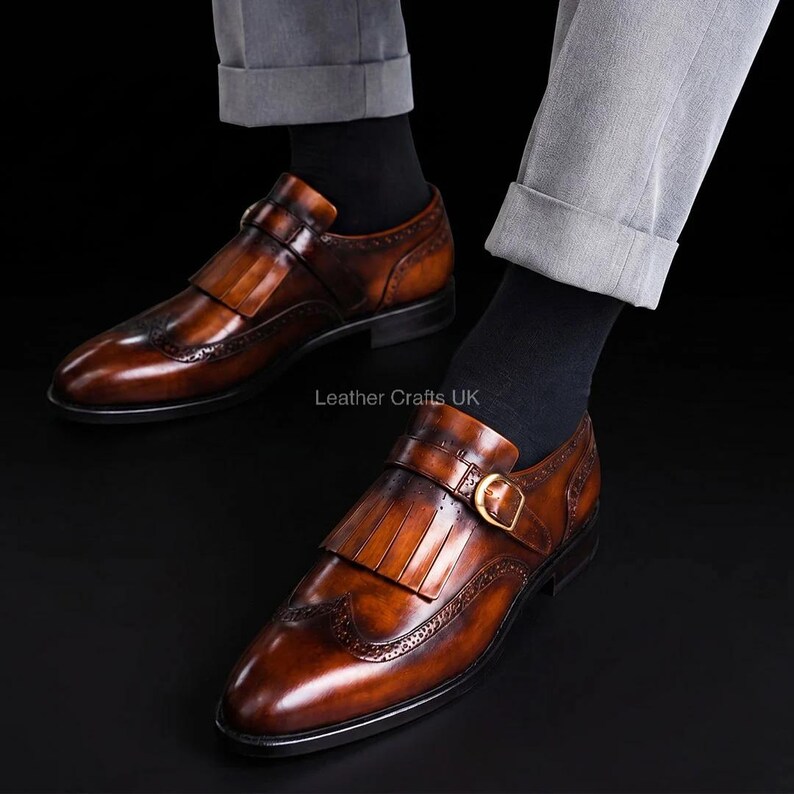 May include: A pair of burnished tan leather monk strap shoes featuring a kiltie fringe detail, a gold-tone buckle, and decorative brogue perforations along the toe cap. The shoes are worn with black socks and light gray trousers. Text reads Leather Crafts UK.
