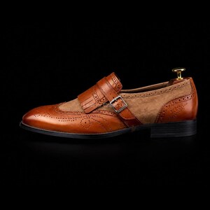 May include: A tan leather monk strap shoe featuring brogue detailing, a kiltie fringe overlay, and a silver-toned metal buckle. The shoe has a smooth leather toe cap and heel counter with a textured suede side panel, set on a black sole.