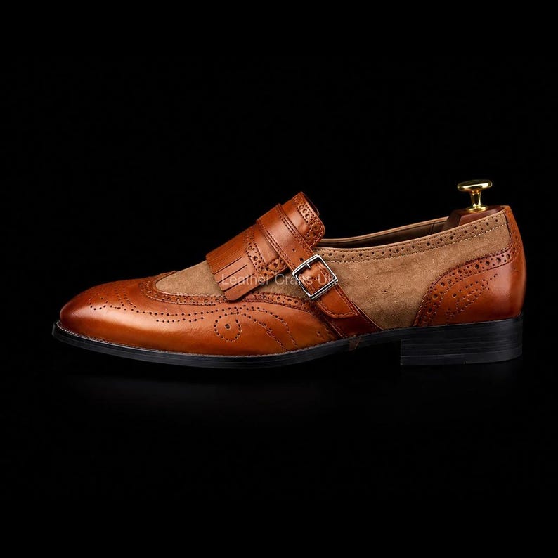 May include: A tan leather monk strap shoe featuring brogue detailing, a kiltie fringe overlay, and a silver-toned metal buckle. The shoe has a smooth leather toe cap and heel counter with a textured suede side panel, set on a black sole.