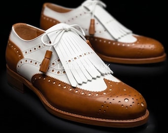 May include: A pair of two-tone wingtip oxford shoes in white and tan leather. The shoes feature decorative brogue perforations, a white leather kiltie fringe over the tongue, white laces with tan tassels, and a low stacked heel on a leather sole.