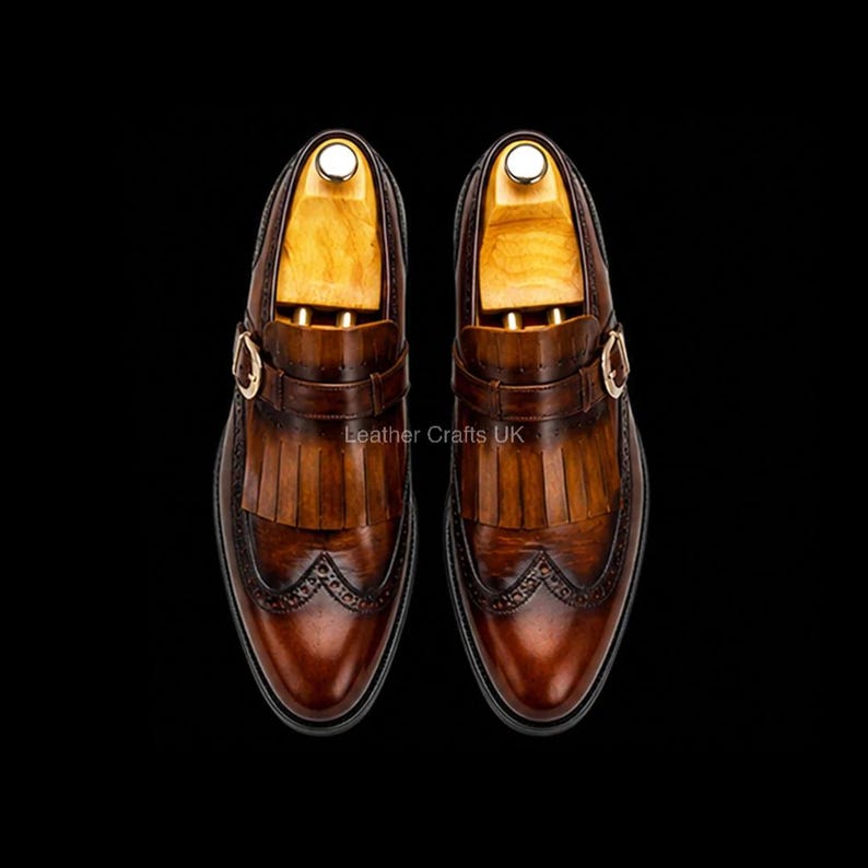 May include: A pair of brown leather monk strap shoes featuring a kiltie fringe, wingtip brogue detailing, and a gold-tone buckle. Each shoe contains a light-colored wooden shoe tree insert. The text Leather Crafts UK is visible in the center.