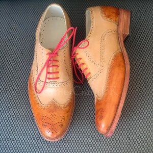 May include: A pair of brown leather brogue shoes with red laces. The shoes have a wingtip design and are made from a combination of light and dark brown leather.