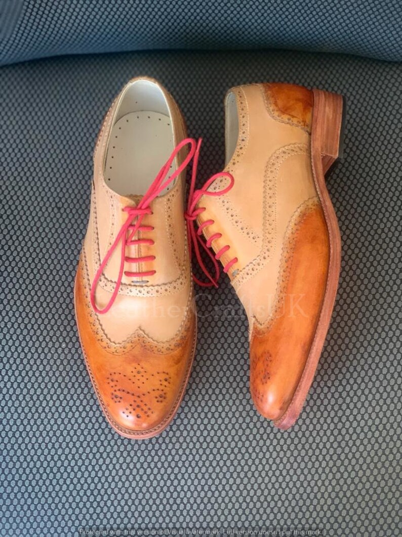 May include: A pair of brown leather brogue shoes with red laces. The shoes have a wingtip design and are made from a combination of light and dark brown leather.