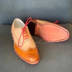 May include: A pair of brown leather Oxford shoes with a wingtip design. The shoes have a red lace and a light brown leather upper. The shoes are on a grey textured surface.