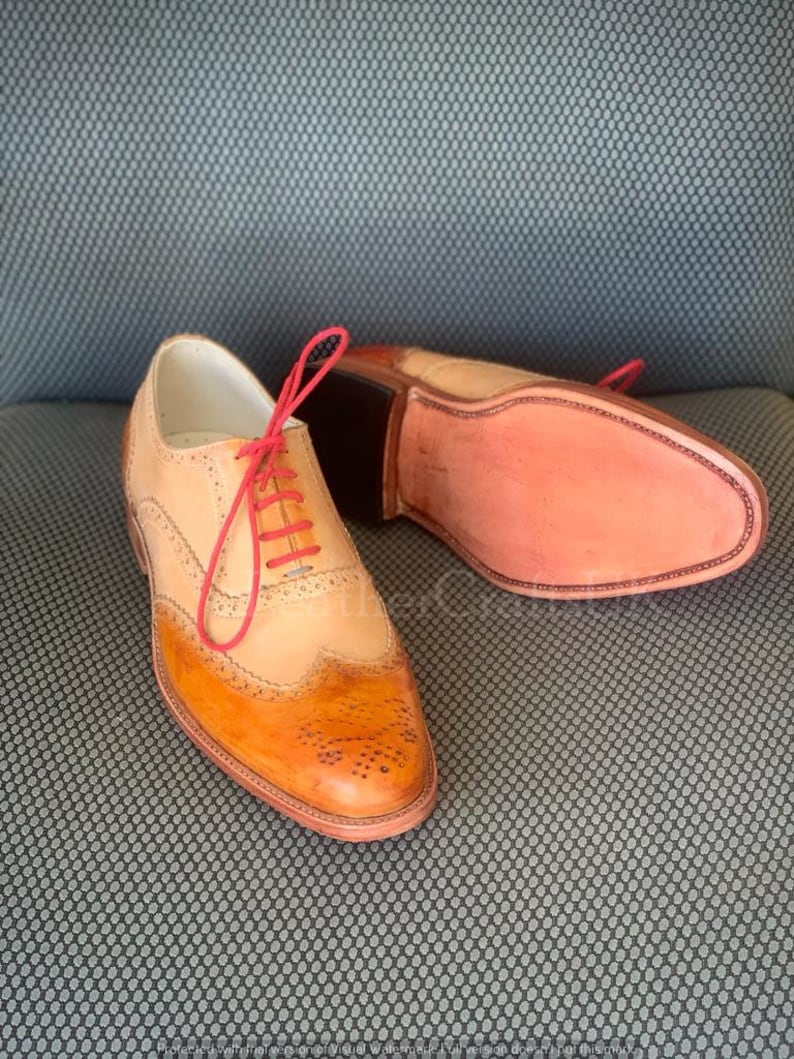 May include: A pair of brown leather Oxford shoes with a wingtip design. The shoes have a red lace and a light brown leather upper. The shoes are on a grey textured surface.