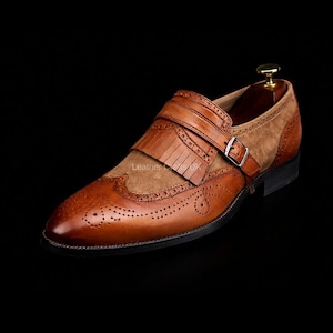 May include: A tan and brown leather monk strap shoe featuring a kiltie fringe, brogue detailing, and a silver-toned metal buckle. The shoe has a smooth leather toe cap with decorative perforations and a suede-like textured side panel.