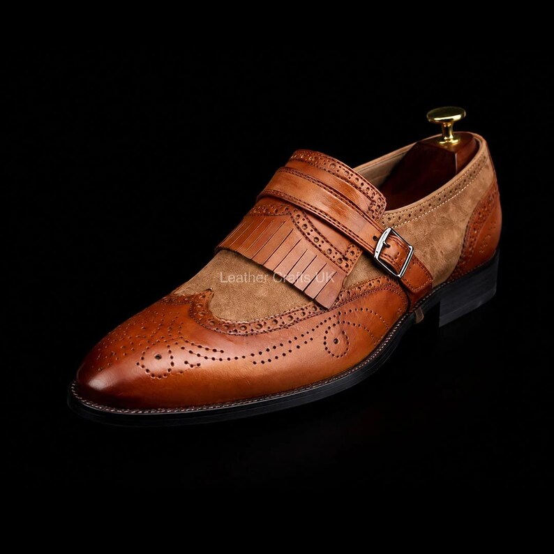 May include: A tan and brown leather monk strap shoe featuring a kiltie fringe, brogue detailing, and a silver-toned metal buckle. The shoe has a smooth leather toe cap with decorative perforations and a suede-like textured side panel.