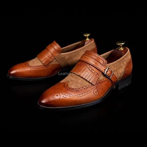 May include: A pair of tan leather and suede monk strap shoes featuring a kiltie fringe detail, brogue perforations on the toe cap, and a silver-toned metal buckle closure. The shoes are set on a dark sole and include gold-toned shoe trees.