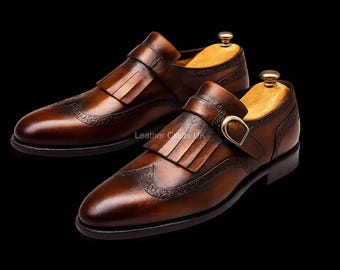 May include: A pair of brown leather monk strap shoes featuring a kiltie fringe detail, a gold-toned buckle, and decorative brogue-style perforations along the toe cap. The shoes are fitted with light-colored wooden shoe trees inside.
