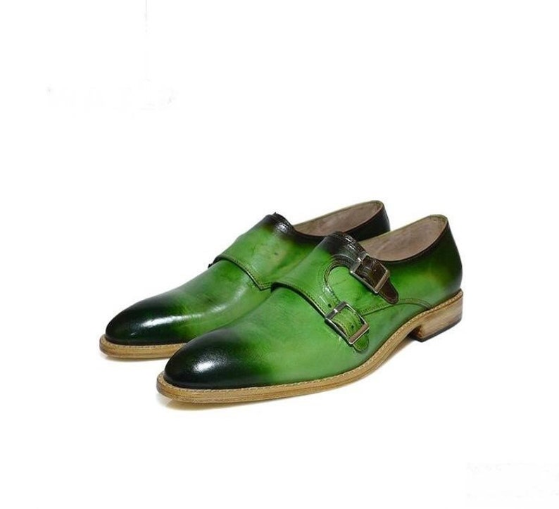 May include: A pair of green leather dress shoes with double monk straps. The shoes have a brown leather sole and a pointed toe.