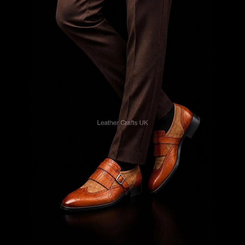 May include: A pair of tan leather monk strap shoes featuring a kiltie fringe detail, brogue perforations, and a silver-toned buckle closure. The shoes are worn with dark brown trousers against a black background. Text reads Leather Crafts UK.
